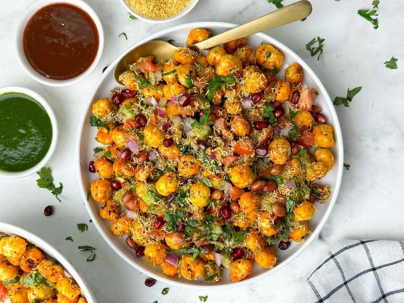 A bowl of makhana bhel topped with sev, pomegranate, tomato, onion and coriander, with green and tamarind chutneys alongside