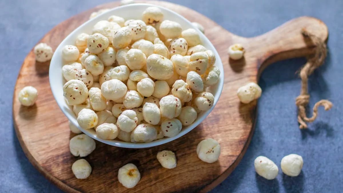 A white bowl of plain roasted makhana on a round wooden board, with loose fox nuts scattered around it