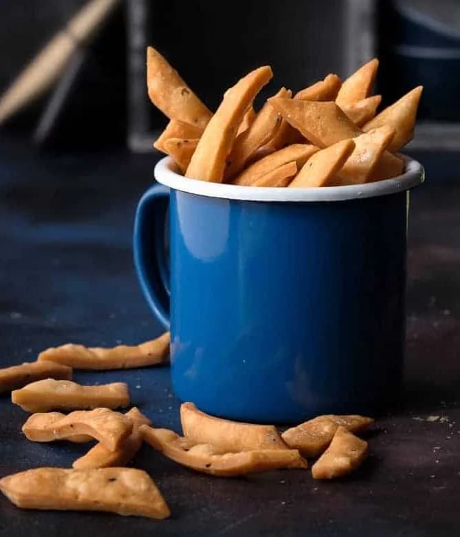 Crispy Namak Pare piled in a blue enamel mug with more pieces scattered on a dark surface