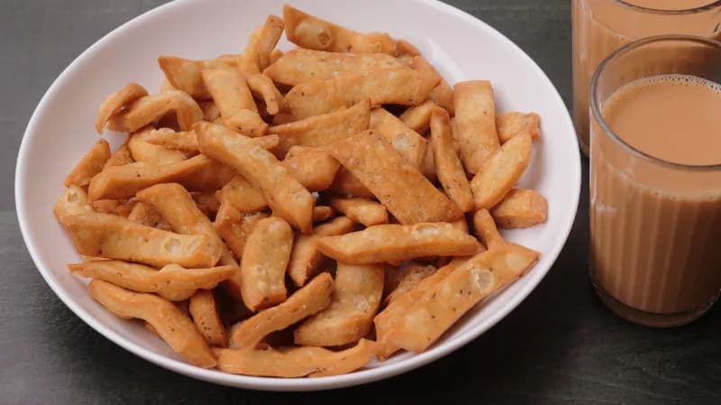 A white bowl of crispy Namak Pare beside two glasses of masala chai, a classic Indian tea time snack pairing