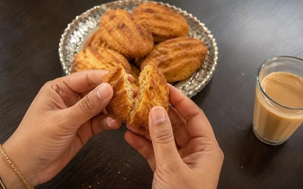 Hands breaking a piece of Thekua beside a glass of chai